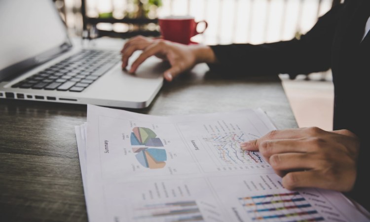 Manos analizan gráficos en papel, junto a un portátil sobre una mesa, con una taza roja al fondo, destacando análisis de datos y productividad.