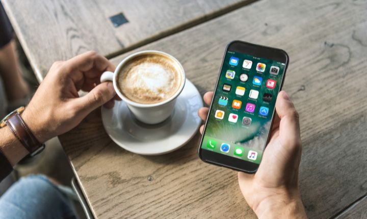 Una mano sostiene un smartphone mostrando aplicaciones, mientras otra mano sujeta una taza de café en una mesa de madera. Ideal para marketing digital.