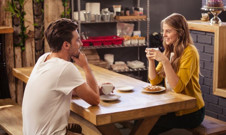 Personas sonriendo, tomando café en una mesa de madera, en un acogedor café con estantes llenos de utensilios detrás. Conversación relajada y amistosa.
