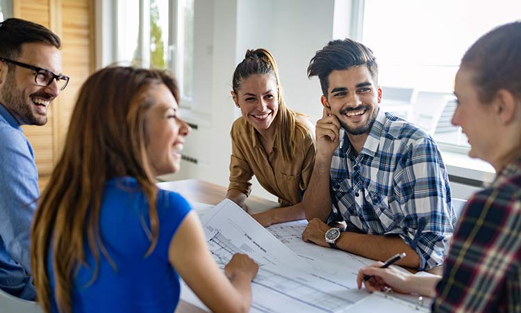 Grupo joven sonriente colaborando en un proyecto, sentados alrededor de una mesa con planos, en una oficina luminosa. Marketing, trabajo en equipo.