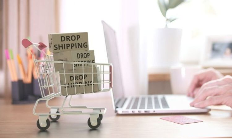 Carrito pequeño con cajas "DROP SHIPPING" en un escritorio; al fondo, manos usando portátil. Ambiente de oficina, enfoque en comercio electrónico.