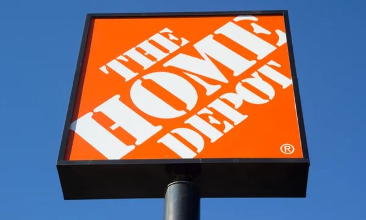 Cartel cuadrado naranja con texto: "THE HOME DEPOT" en letras blancas mayúsculas, sobre un poste negro, y cielo azul como fondo. Ideal para marketing.