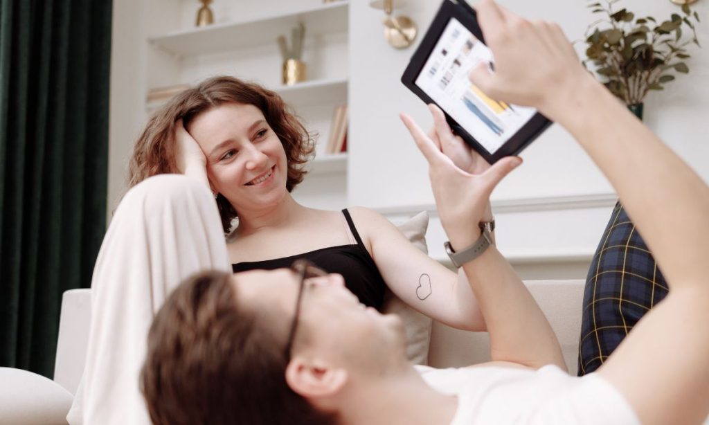 The image shows a couple in a relaxed environment, possibly in their living room. The woman is sitting on the sofa, looking at the man who is lying down and holding a tablet. He seems to be showing her something on the screen while she smiles. The scene conveys a sense of comfort and closeness.