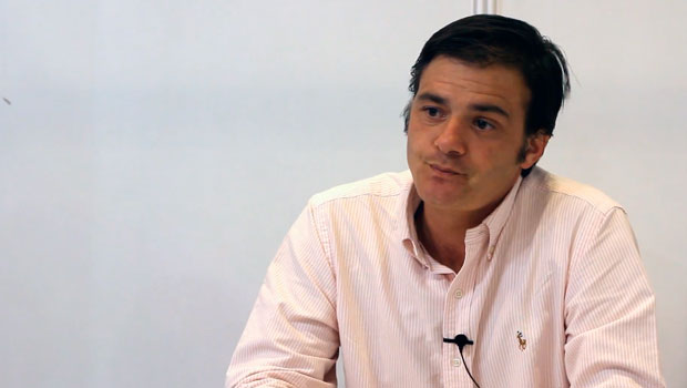 Un hombre lleva una camisa clara, parece estar reflexionando mientras está sentado y mirando al frente; fondo minimalista blanco.