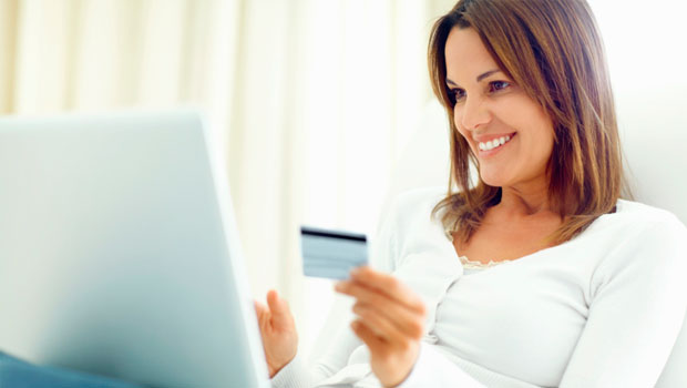 Mujer sonriendo usa portátil y sostiene tarjeta de crédito; ambiente cómodo en casa. Palabras clave: compra online, comodidad, ecommerce, usuario satisfecho.