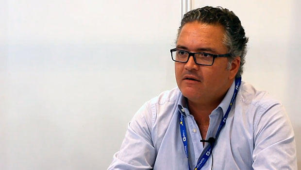 Hombre con camisa blanca, sentado y hablando. Usa gafas y una cuerda azul con tarjetas colgada al cuello. Fondo blanco. Marketing. Negocios.