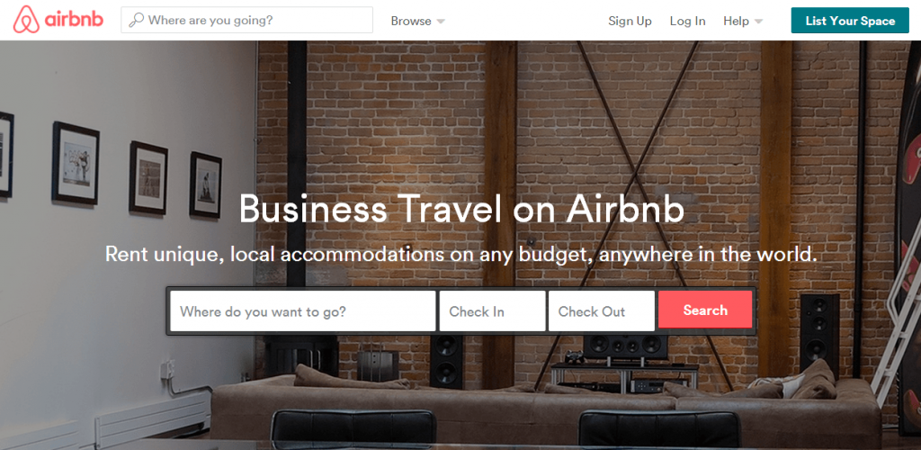 Pantalla de Airbnb mostrando búsqueda de alojamientos. Texto: "Business Travel on Airbnb. Rent unique, local accommodations on any budget, anywhere in the world." Ambiente hogareño con sofá y pared de ladrillos.