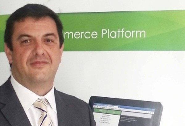 Hombre sonriente, viste traje elegante; frente a un cartel que dice "Commerce Platform", al lado de una pantalla en un entorno profesional.