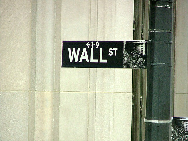 Señal indica "WALL ST" con flecha a la izquierda, fijada en un poste urbano. Fondo de edificio. Texto sugiere ubicación financiera icónica.