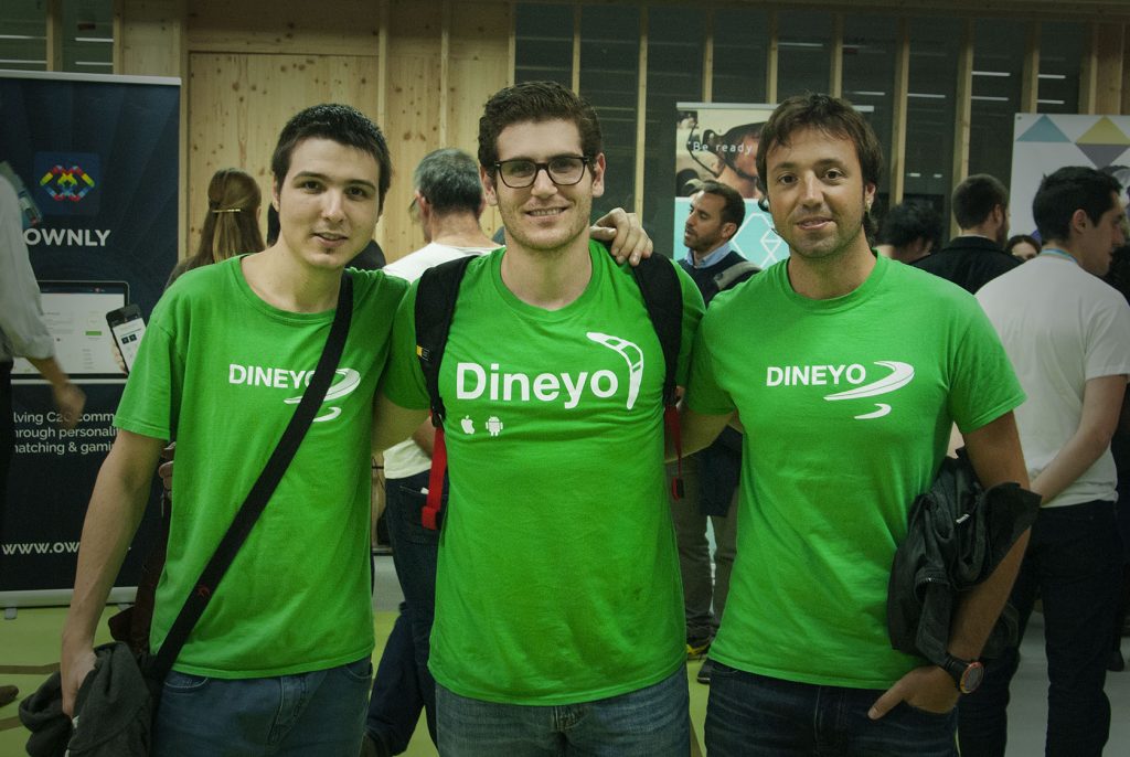 Tres personas sonrientes, con camisetas verdes que dicen "Dineyo," se abrazan en un evento concurrido. Están en un entorno de networking.
