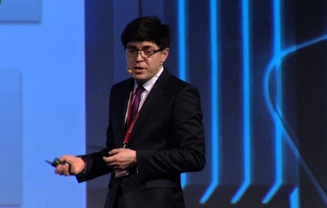 Hombre hablando en un escenario, sujetando un mando. Fondo con luces azules y diseño geométrico. Contexto de conferencia o presentación de negocio.
