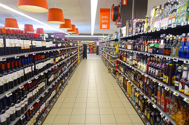 Pasillo de supermercado con estantes llenos de botellas de vino y licor; letrero rojo en el centro que dice "Info prix". Ambiente comercial ordenado.