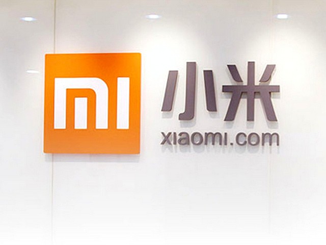 Logotipo de Xiaomi, letras chinas y sitio web, en una pared blanca iluminada; enfoque en tecnología y diseño moderno.