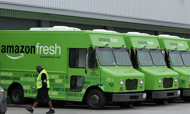 Camión verde de Amazon Fresh estacionado, empleado caminando enfrente; entorno de almacén. Texto: "amazonfresh", "amazonfresh.com", palabras sobre productos.