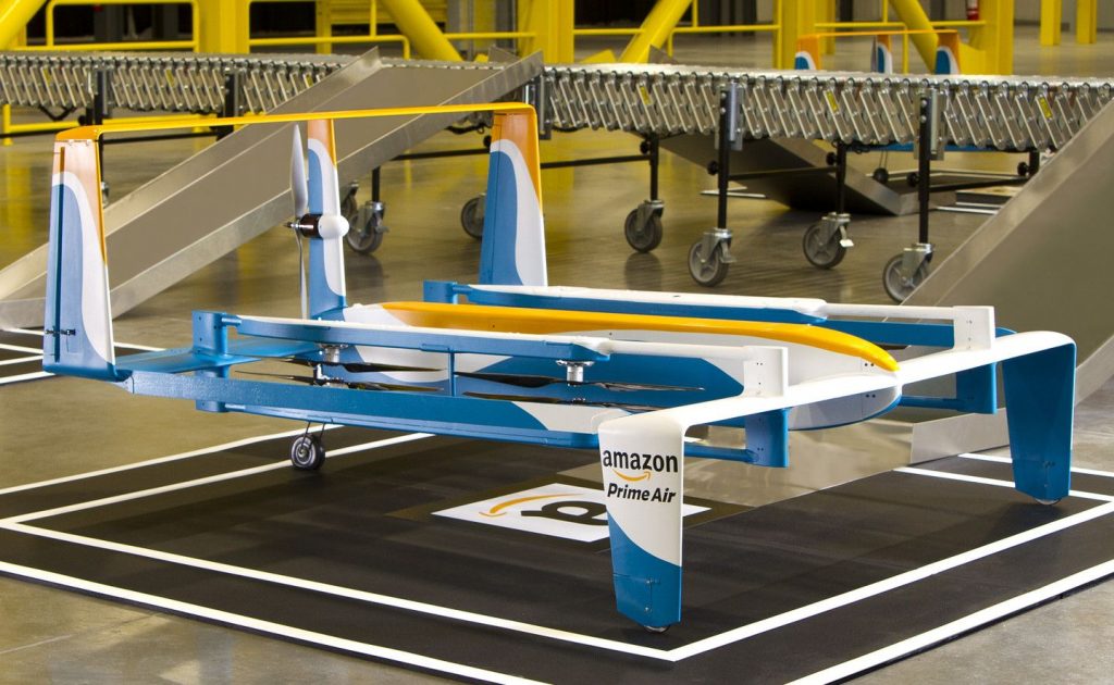 Dron colorido de "Amazon Prime Air" estacionado en almacén moderno con rampas y ruedas, listo para entregas rápidas y eficientes. Tecnología avanzada.