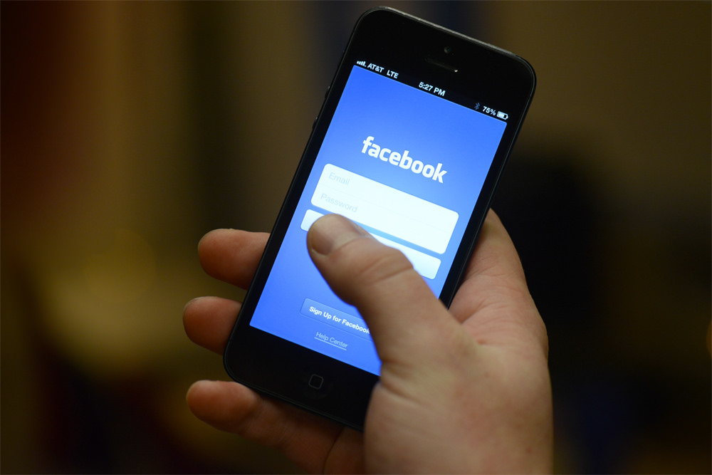 Mano sosteniendo un móvil con pantalla mostrando la página de inicio de sesión de Facebook. Texto: "facebook, Email, Password, Sign Up for Facebook, Help Center". Contexto de uso digital y conexión.
