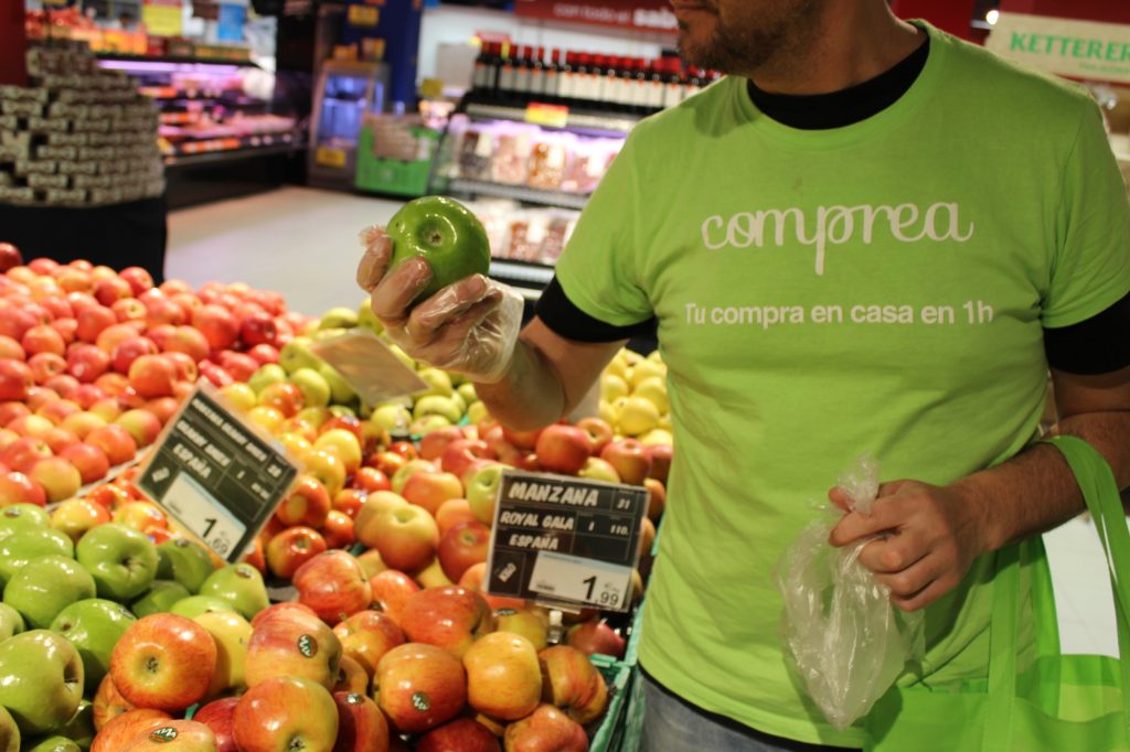 Persona observando manzana mientras compra en supermercado. Lleva camiseta verde con texto "compra en casa en 1h". Frutas variadas alrededor.