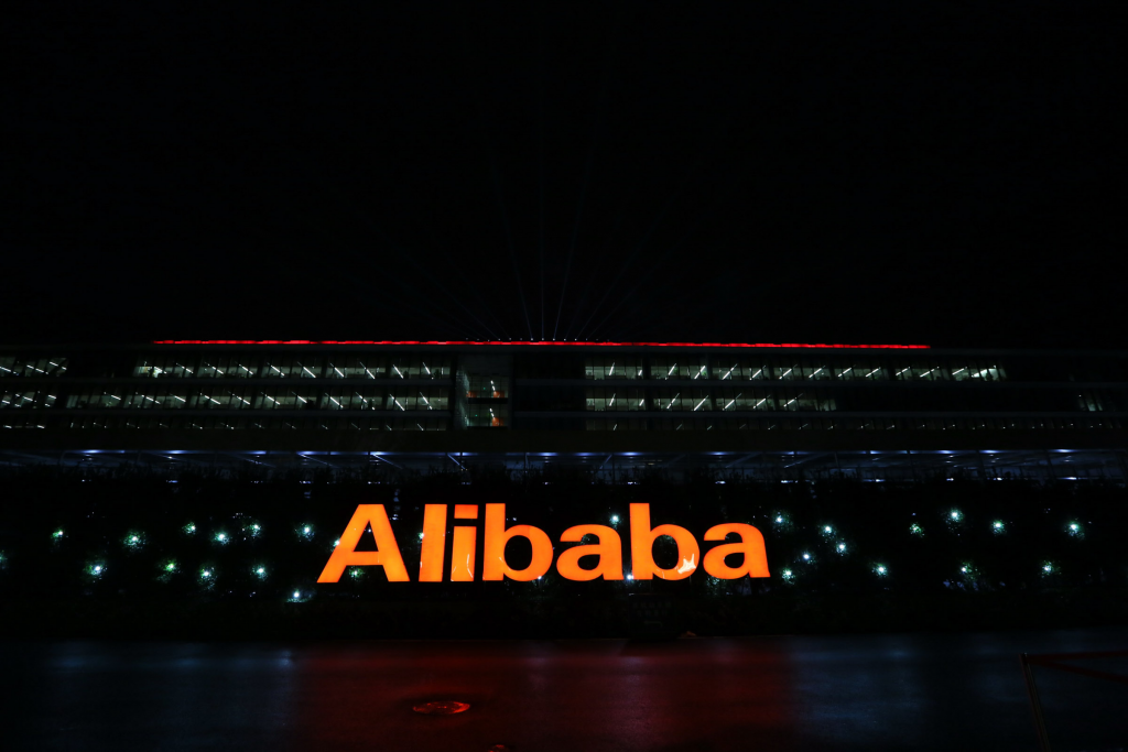 Texto "Alibaba" iluminado en naranja frente a edificio con luces rojas en el techo, ambiente nocturno. Palabras clave: marca, tecnología, innovación.
