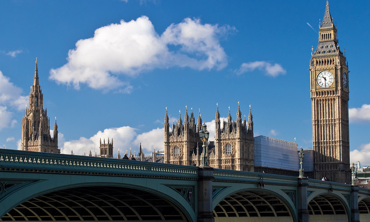 Reloj alto sobre torre icónica destaca bajo cielo azul, junto a arquitectura gótica, cruzando puente verde, alusión clásica y turística británica.