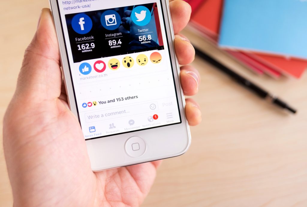 Teléfono muestra redes sociales y emojis; mano lo sostiene sobre escritorio con bolígrafo y cuadernos. Texto visible: "Facebook 162.9 millions, Instagram 89.4 millions, Twitter 56.8 millions".
