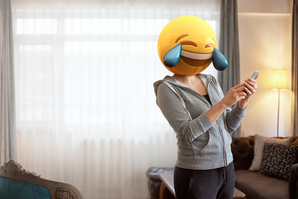 Persona con emoji de risa en la cabeza, usando un móvil en una sala de estar iluminada. Ideal para campañas de humor y tecnología.
