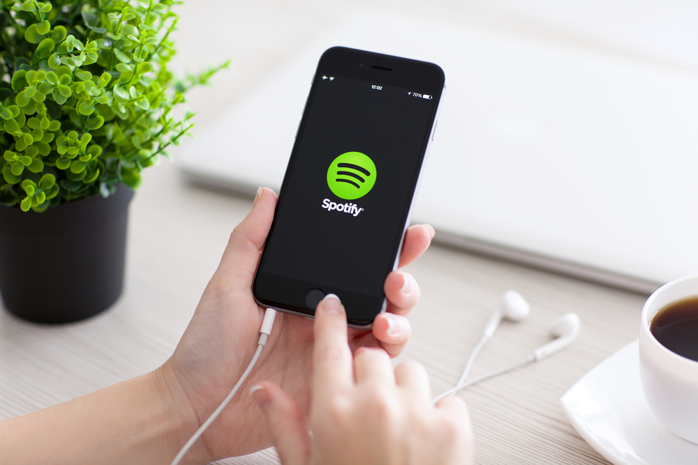 Teléfono muestra Spotify activo; manos sostienen dispositivo. Contexto: escritorio claro con portátil, auriculares, taza de café y planta. Optimiza tu experiencia musical.