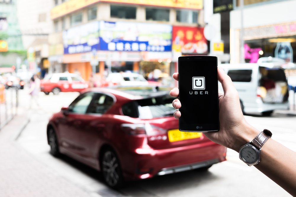 Mano sostiene un móvil mostrando la app de Uber, parado frente a un coche rojo en una calle animada de la ciudad. Ideal para marketing urbano.