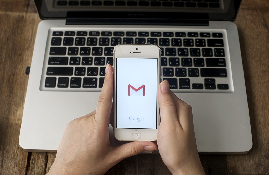 Manos sostienen móvil con logo de Gmail en pantalla, sobre portátil abierto. Texto visible: "Google". Ambiente oficina, tecnología, comunicación digital.