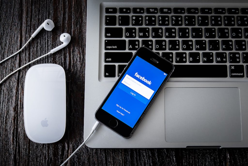 Teléfono muestra pantalla de inicio de sesión de Facebook sobre teclado de laptop. Al lado, ratón y auriculares. Contexto: tecnología y redes sociales.