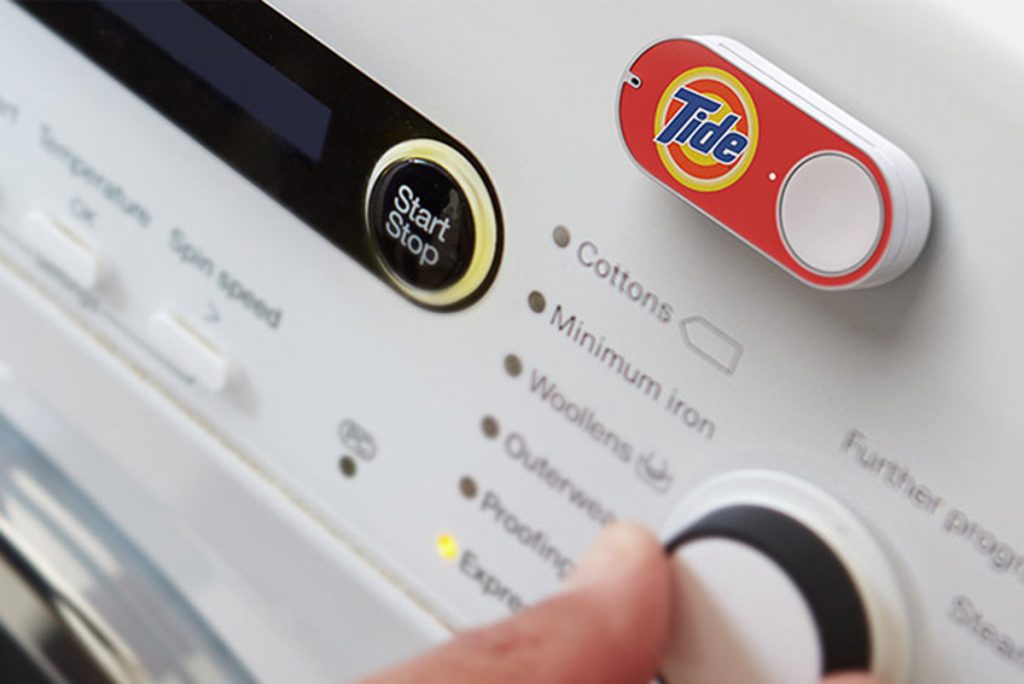 Botón Amazon Dash, con logotipo Tide, colocado en lavadora. Un dedo está a punto de presionarlo, destacando comodidad y rapidez en compras.