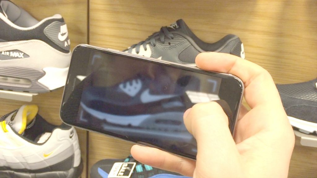 Teléfono enfocado en un zapato deportivo, mientras alguien lo sostiene, en una tienda de calzado moderno. Texto: "AIR MAX", "SALE EX28".
