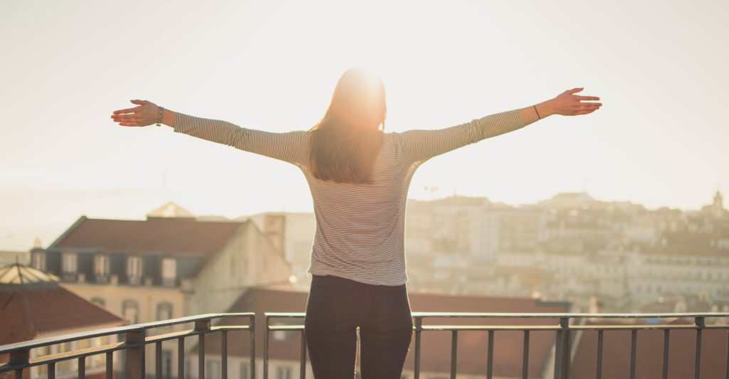 Persona extiende los brazos en un balcón, disfrutando el sol de la mañana. Al fondo, se percibe una ciudad bajo la luz dorada. Inspiración y libertad.