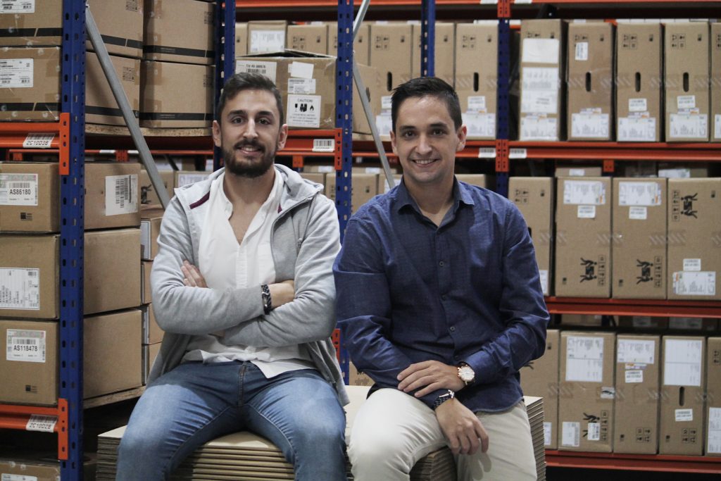 Dos hombres sentados sonríen frente a estanterías de cartón en un almacén, reflejando logística y gestión de inventario eficaces.