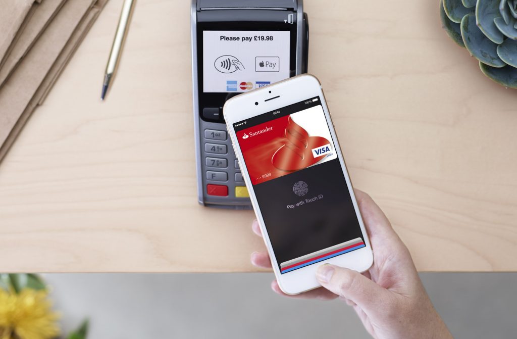Teléfono mostrando pago con tarjeta Visa a punto de realizar una transacción sin contacto en un datáfono; mensaje: "Please pay £19.98". Contexto de mesa.