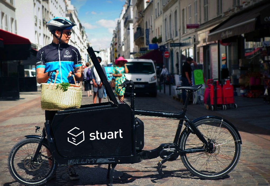 Repartidor en bicicleta de carga con logo "stuart", sostiene cesta de compras en calle urbana. Entorno comercial vibrante. Ideal para entregas rápidas.