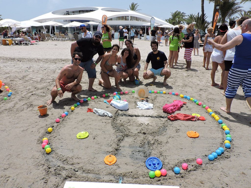 Grupo de personas sonríe posando en la arena. Están rodeados de pelotas de colores y objetos de playa. Al fondo, edificio moderno y palmeras. Ideal para promoción de eventos al aire libre y actividades en la playa.