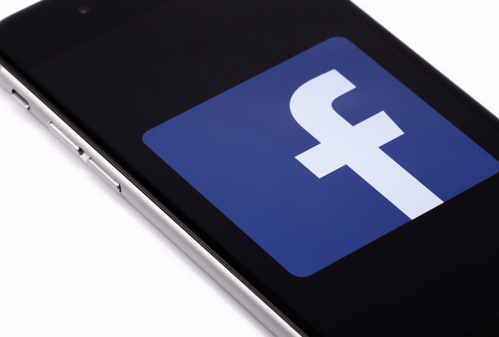 Teléfono mostrando el logotipo de Facebook en pantalla; en un fondo negro, símbolo central en azul y blanco; comunicación digital, redes sociales.