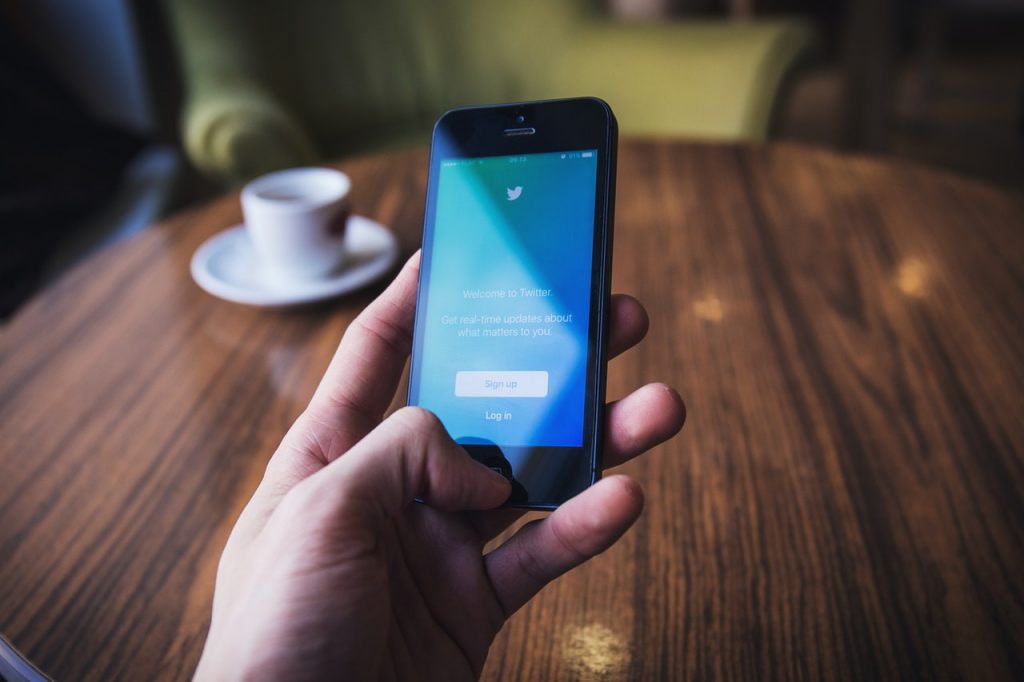 Teléfono muestra pantalla de inicio de Twitter; mano sostiene el dispositivo. Fondo: mesa de madera con taza de café. Texto: "Welcome to Twitter. Get real-time updates about what matters to you. Sign up. Log in". Conectar digitalmente, redes sociales.