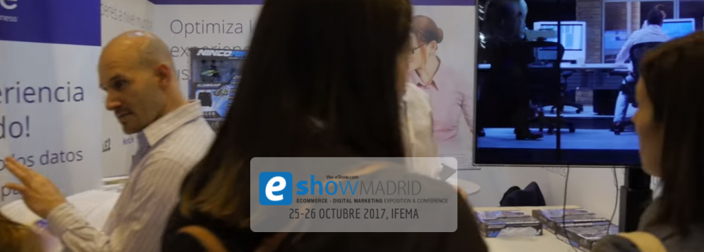 Persona hablando a un grupo en una feria sobre marketing digital en Madrid. Carteles promocionales y pantalla de fondo. Texto: "the-eShow.com Madrid eCommerce - Digital Marketing Exposition & Conference 25-26 octubre 2017, IFEMA."
