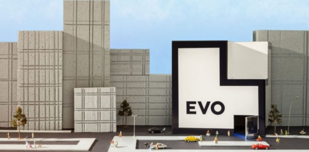 Edificio de banco Evo, fachada blanca geométrica moderna. Personas y coches en miniatura transitan frente al edificio en una ciudad con rascacielos.