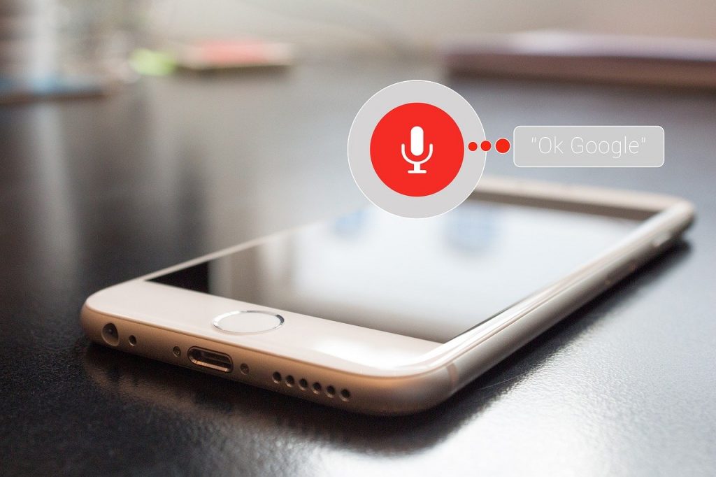Teléfono inteligente en mesa, pantalla activa con el icono del micrófono rojo para el asistente de voz "Ok Google". Innovación tecnológica.