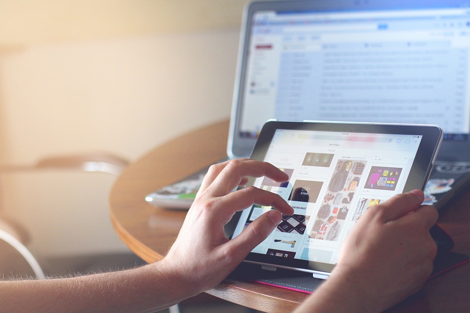Manos utilizando tableta, navegando imágenes; portátil con pantalla de correo en segundo plano sobre mesa de madera. Tecnología digital en oficina.