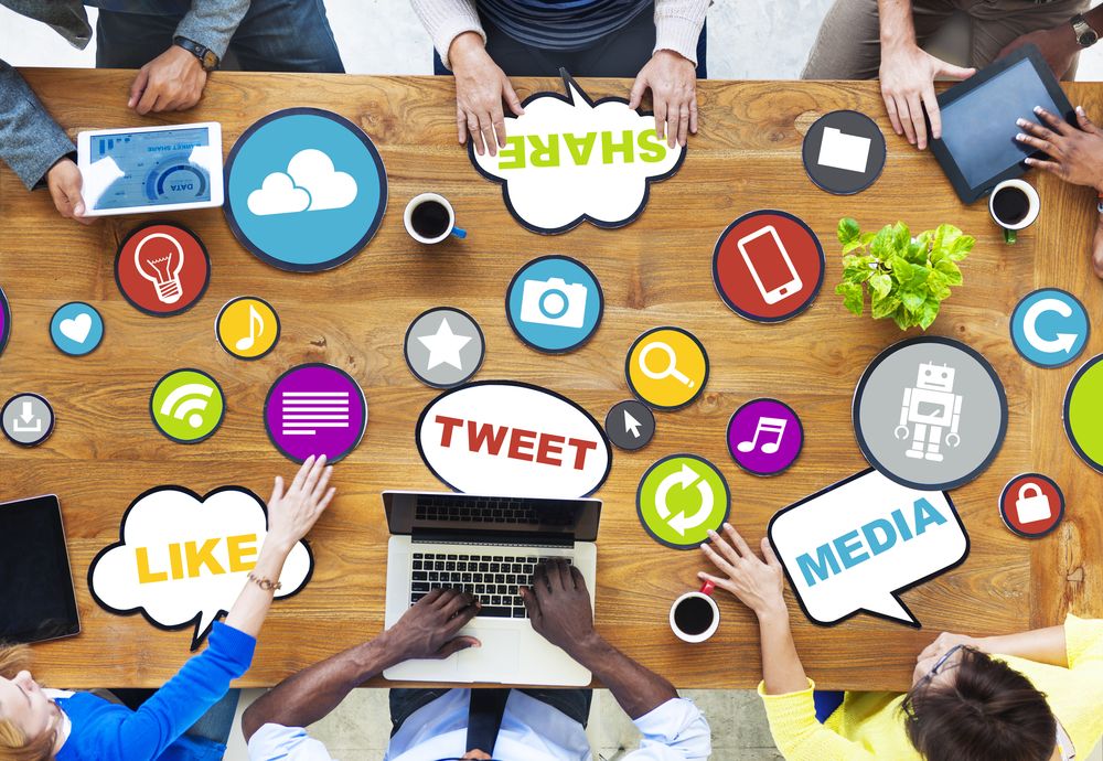 Personas trabajando entorno a una mesa de madera con iconos de redes sociales. Textos: “LIKE”, “SHARE”, “TWEET”, “MEDIA”. Ambiente colaborativo digital.