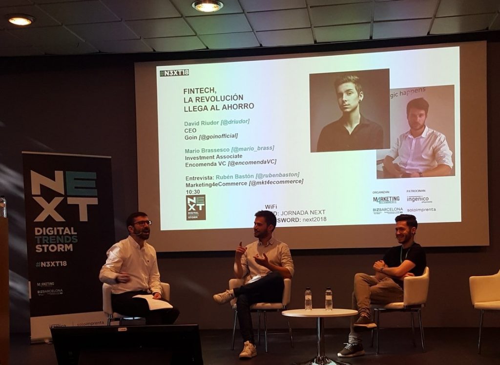 Tres hombres sentados en un escenario participan en una charla sobre "Fintech, la revolución llega al ahorro" en el evento NEXT Digital Trends, INTERACTÚAN en una sala de conferencias. Texto: "N3XT18, David Riudor, Mario Brassesco, entrevista: Rubén Bastón, Marketing4eCommerce. WiFi: Jornada NEXT, password: next2018".