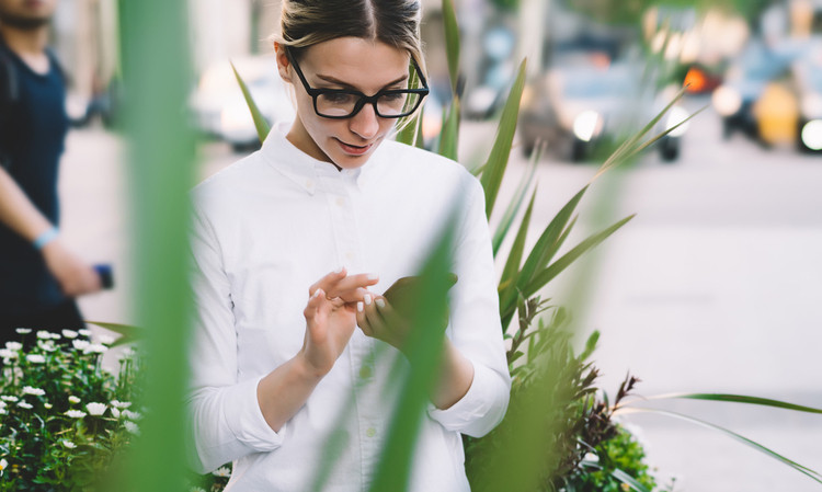 Persona con gafas revisa su móvil mientras camina por la ciudad; plantas y luz diurna en el entorno. Tecnología y estilo urbano.