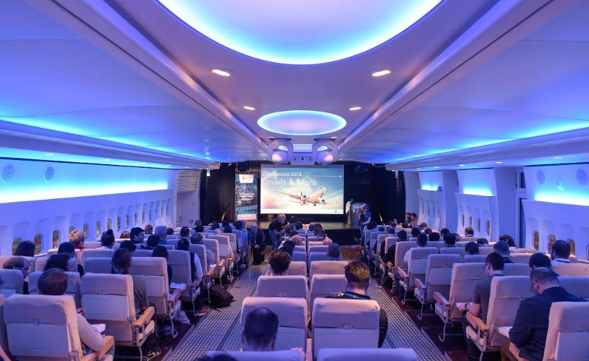 Pasajeros-asistentes sentados en silla de avi&oacute;n escuchan una presentaci&oacute;n en pantalla grande. Entorno simula interior de avi&oacute;n comercial. Ideal para eventos corporativos.