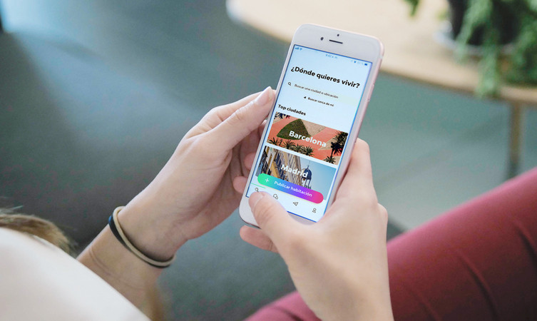 Manos sostienen un teléfono mostrando una app de búsqueda de viviendas. Texto: "¿Dónde quieres vivir? Top ciudades: Barcelona, Madrid." Ambiente relajado.