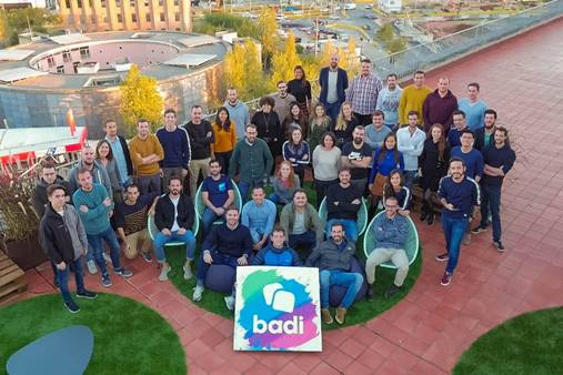 Un grupo de personas posando juntas sobre una terraza con suelo de baldosa roja y c&eacute;sped artificial; en el centro, un cartel con el texto "badi". Ambiente corporativo, trabajo en equipo.