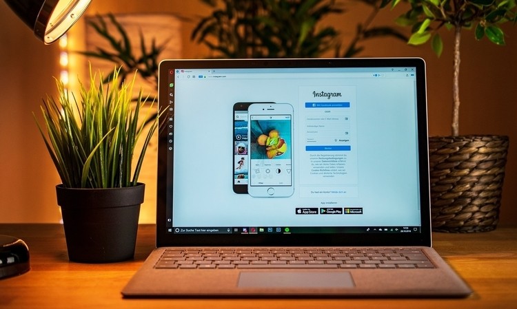 Portátil muestra página de inicio de Instagram; rodeado de plantas en oficina acogedora. Texto en pantalla: "Instagram" y campos de inicio de sesión. Marketing digital.