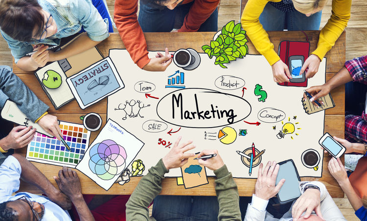 Mesa llena de papeles de marketing con gráficos y dibujos, rodeada de personas colaborando; destacan móviles, café y una planta. Texto: "Marketing".